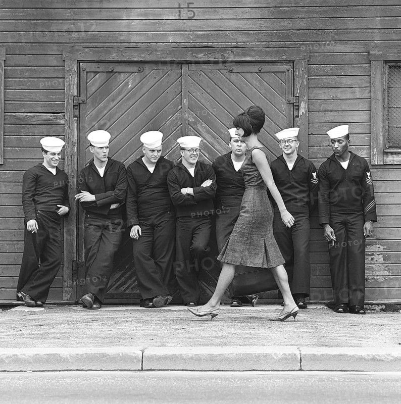 Fashion in the 1960s. A group of American sailors are standing at the docks on shore leave and makes a nice addition to the fashion shoot. A young model passes them wearing a dress with matching handbag and shoes. Sweden 1964 ref BV59-5