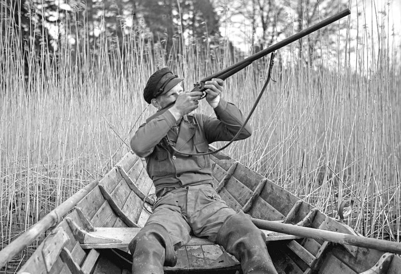 In the 1930s. A man sits in a rowing boat and aims his rifle upwards to shoot ducks. Sweden 1939. Kristoffersson ref 155-11