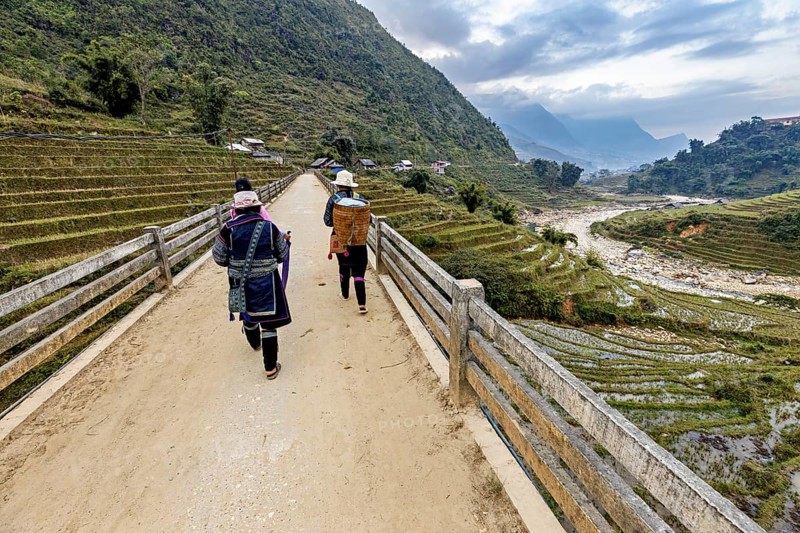Two people walk on a bridge with a view of terraced rice paddies and mountains, local women farmers walk through the countryside near Sa Pa in Vietnam