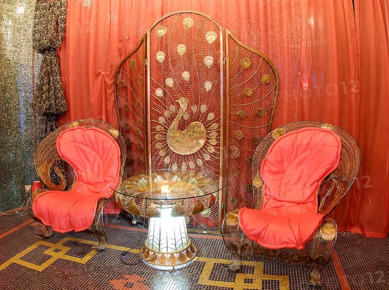 Nice metal delicate forged furniture in oriental style, peacock theme prevailing: two armchairs, glass coffee table with lighting inside its stand and folding screen against red orange curtains