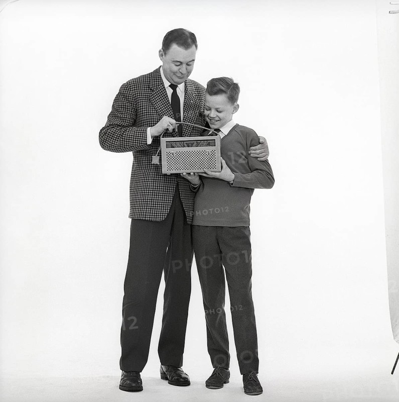 In the 1960s. Father and son with a radio. It's a typical 1960s radio with a cover of perforated leather. Battery operated transistor radio with AM and FM bands. The radiostations and frequenses are displayed on the front, and a knob to turn and adjust to get best possible reception. Sweden 1960s CX88-1