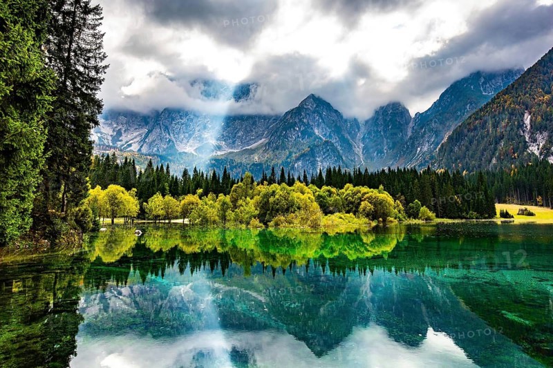 The sun's rays break through the clouds. The smooth surface of the water reflects forest and mountains. Gorgeous reflections. The lake Fusine in Northern Italy. Mountains covered in mist
