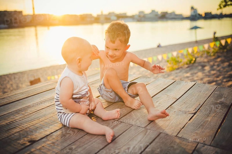 Two little boys sitting on the pier on the river bank. Concept of friendship and fraternity. Sunny joyful summer day. Children sitting on pier. Two children of different age sitting on a wooden pier