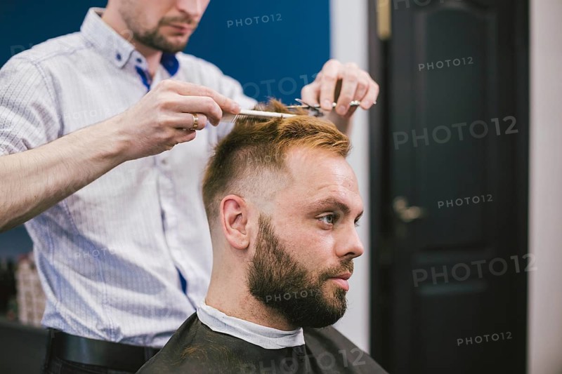Barber Making Haircut Bearded Man In Barbershop. Professional stylist cutting client hair in salon. Barber using scissors and comb. Skillful hairdresser cutting male hair. Hair Care Service Concept