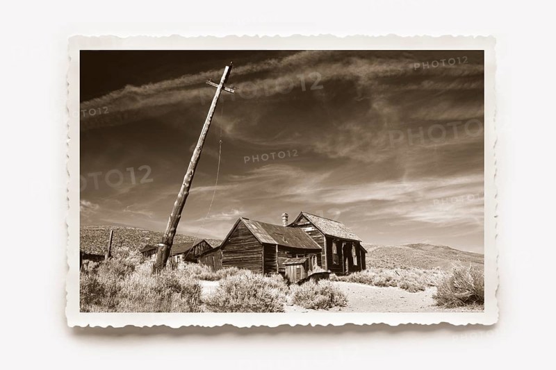 Lonely wooden house in Bodie, in front of it a crooked lamp post. Dry grass and undergrowth in the foreground. Photo in sepia tone with a white jagged edge and a sunburst shadow. Lonely wooden house in Body, besides a crooked lighting mast. In the foreground dry graß and dense undergrowth. Photo in sepia toning with white jagged edges