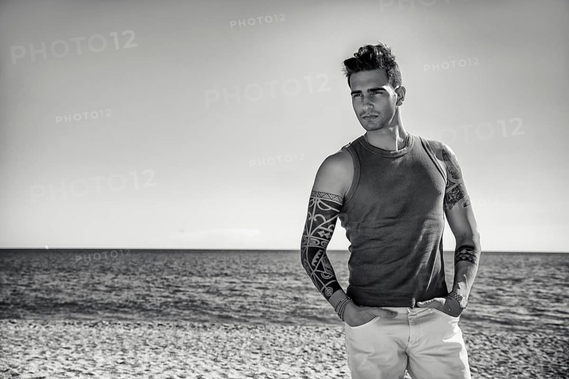Three Quarter Shot of a Handsome Athletic Young Man in Trendy Attire, on a Beach in a Sunny Summer Day, Looking At Camera against Blue Sky Background