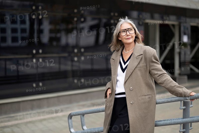An older woman with gray hair and glasses exudes confidence while leaning against a railing in front of a sleek modern building. She wears a chic outfit that showcases her elegant style