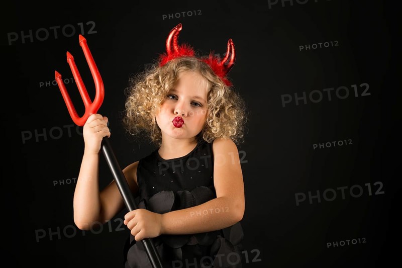 Isolated on black, adorable caucasian blonde little girl in black dress, with red horns on head hold trident both hands, look at camera, serious look