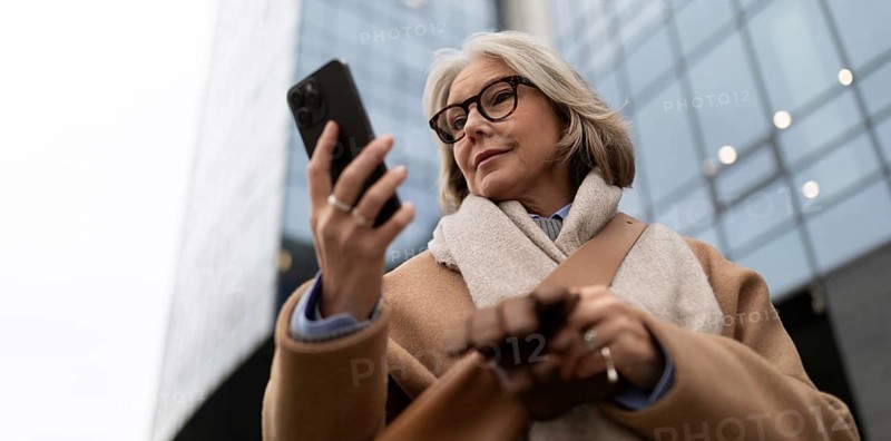 A mature woman wearing glasses and a warm coat is focused on her smartphone, standing on the sidewalk near modern buildings in an urban setting during overcast weather