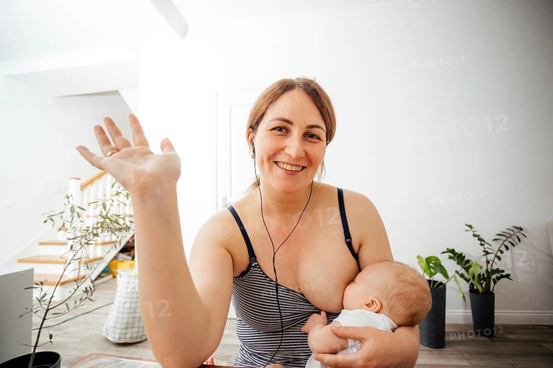 Portrait of cheerful mother in casual top nursing cute newborn while online meeting. Woman combines remote work and raising her child. Webcam view woman looking at the camera