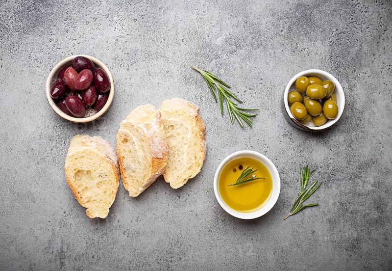 Slices of fresh ciabatta, green and brown olives, olive oil with rosemary, olive tree branches on gray concrete stone rustic background overhead, food photography, food photography