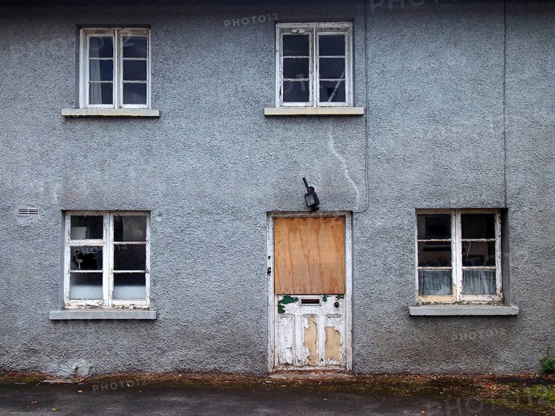 An old abandoned grey derelict house on a residential street with dirty broken windows and a boarded up door