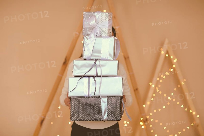 Female holding big stack of presents wrapped in brown paper with white bows on simple background with lights