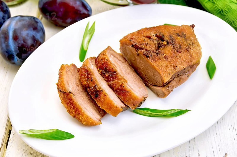 Roasted duck breast with green onions in a white oval plate, towel and plums on a background of light wooden boards