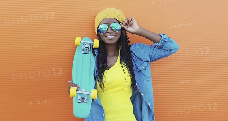 Summer portrait of stylish happy African young woman with skateboard, modern black teenage girl in yellow hat, glasses posing on orange background
