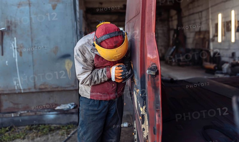 A worker sands a rusty van in an outdoor workshop setting
