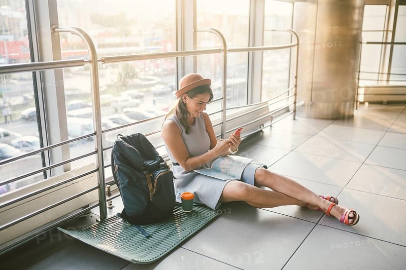 Theme of tourism and travel of young student. Beautiful young caucasian girl in dress and hat sits on floor tourist rug inside terminal airport terminal. Waiting room delayed flight, delay departure