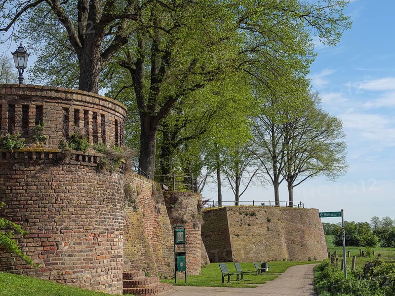 Defence defence tower and brick wall along a walkway, flanked by trees and park benches under a blue sky, beautiful promenade along the Rhine with a tower, lanterns and trees, springtime on the Rhine, Rees, Germany