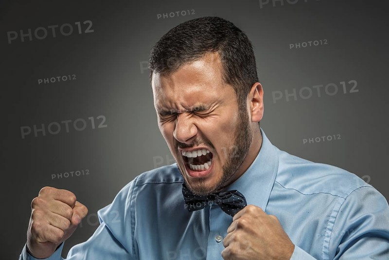 Portrait of angry upset young man in blue shirt and butterfly tie with fists up yelling isolated on gray studio background. Negative human emotion, facial expression. Closeup