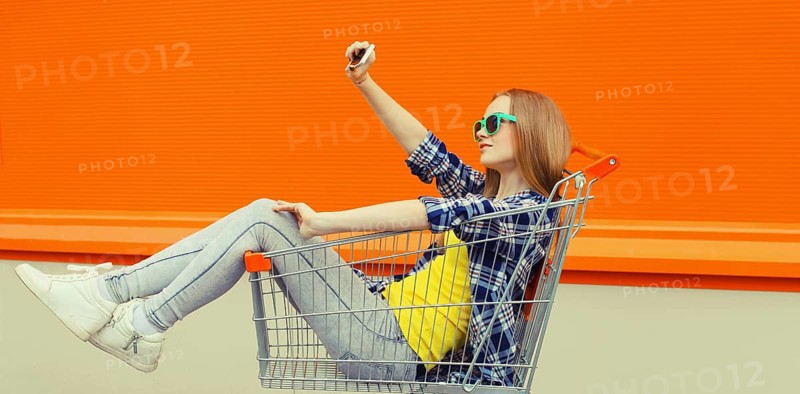 Stylish happy young woman taking selfie with phone, cool modern teenage girl having fun sitting in shopping trolley cart in the city on a summer day