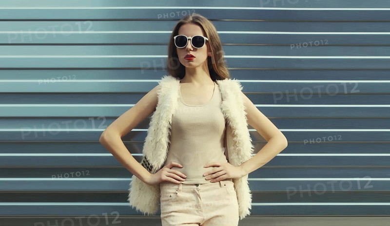 Portrait of modern stylish young woman in white fur vest, glasses, fashionable female model posing on gray silver wall background