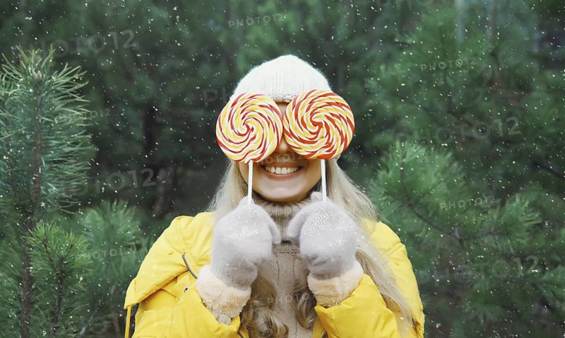Happy funny smiling young woman having fun with lollipop enjoys snow, in winter park against Christmas tree, joyful girl standing in snowy forest with snowflakes, festive background