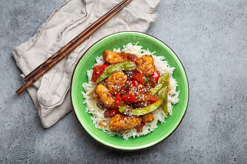 Asian sweet and sour sticky chicken with vegetables stir-fry and rice in ceramic bowl with chopsticks top view on gray rustic stone background, traditional Asian dish, food photography, food photography