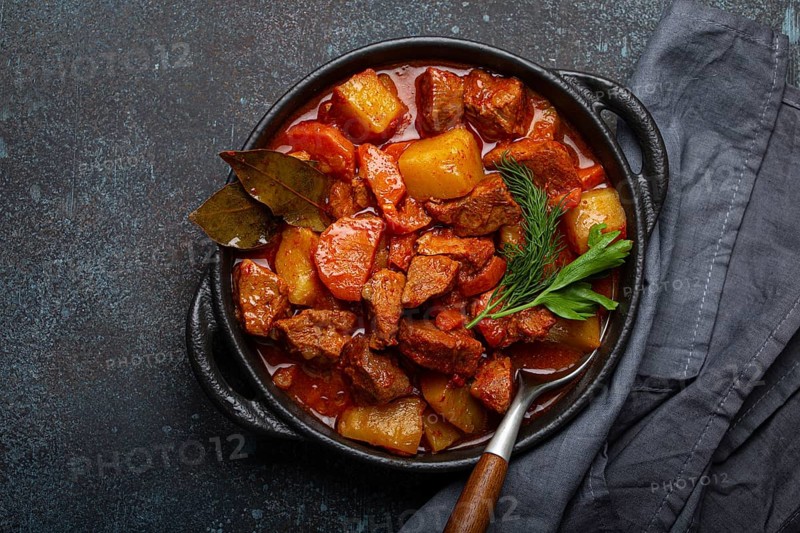Beef meat stew with potatoes, carrot and delicious gravy in black casserole pot with bay leaves and fresh green herbs with spoon on black dark rustic concrete background from above, food photography