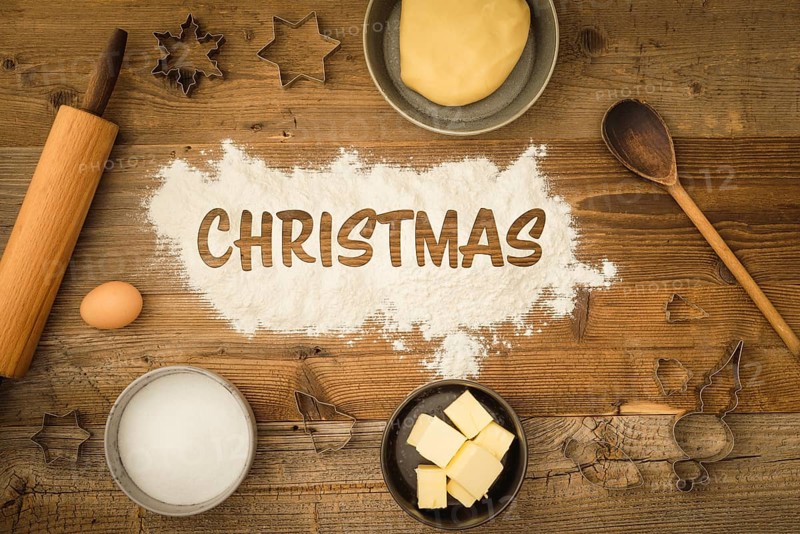 Collection of ingredients and utensils for baking Christmas biscuits with the word Christmas written in flour in the centre of the picture on a dark wooden background