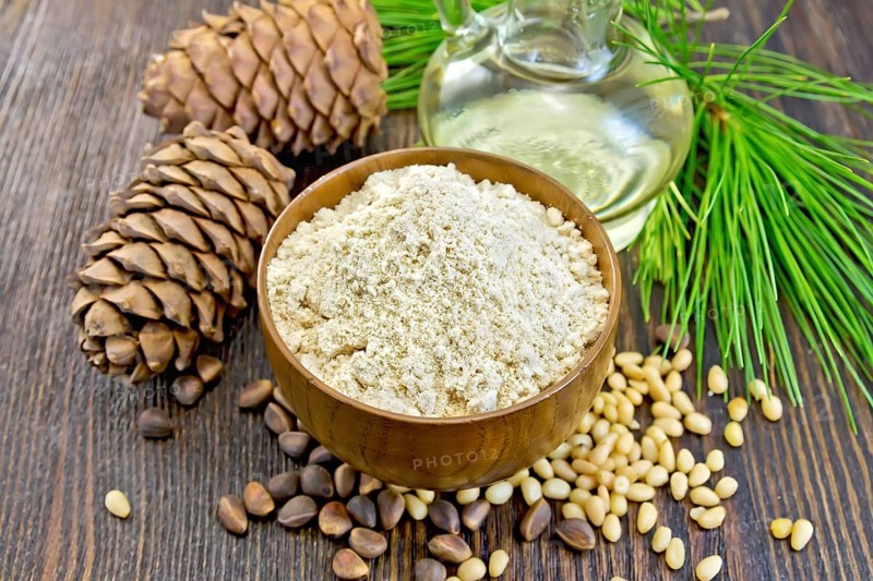 Flour cedar in a wooden bowl, cedar nuts and two cones, the oil in the glass carafe, cedar branch against the backdrop of wooden planks
