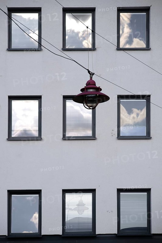 A hanging lamp in front of the façade of a residential building with windows reflecting the clouds
