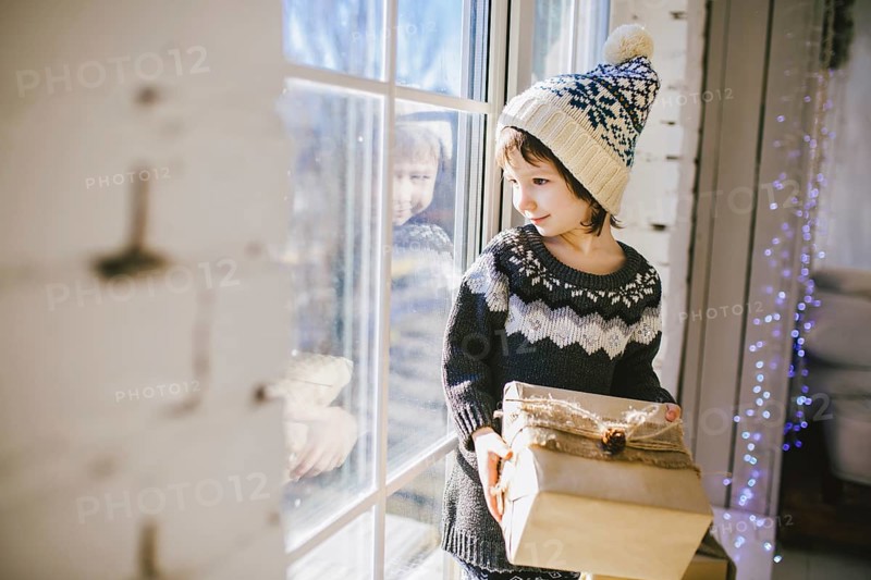 Child sitting at the window on a sunny Christmas day, making presents wrapped in boxes in paper. dressed in knitted warm woollen clothes and hat. Inside the house with a stylish interior
