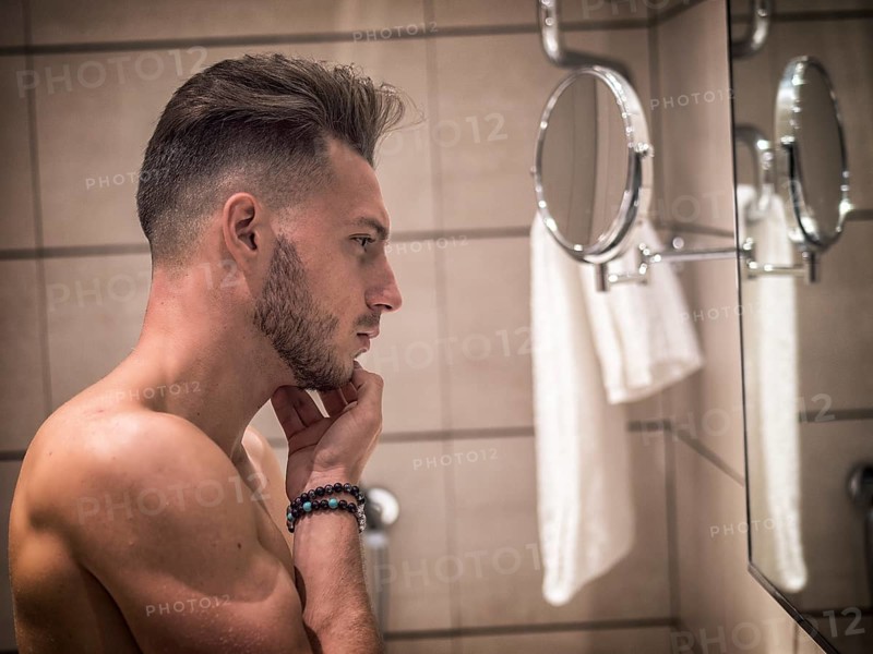 Shirtless muscular handsome young man looking at himself in bathroom mirror in the morning