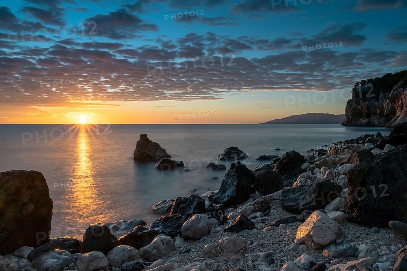 A colorful sunrise at Cala Gonone with black and white rocks and boulders and a sunburst