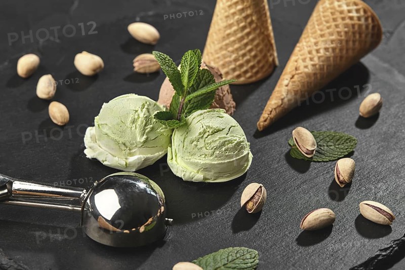 Frozen chocolate and pistachio ice cream decorated with mint, waffle cones with scattered pistachios are nearby, served with a scoop on a stone slate over a black background. Close-up