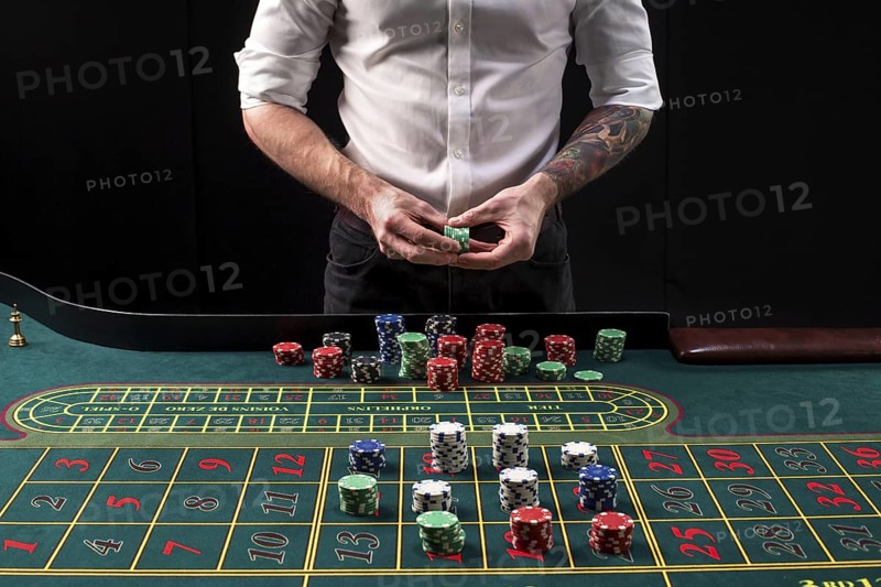 A close-up vibrant image of green casino table with roulette, with the hands of croupier and multicolored chips, on black background. Casino. Gambling. Roulette. Betting