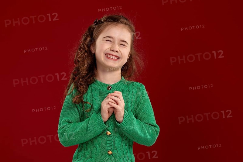 Portrait of a laughing tiny kid with a long, curly hair looking happy. Charming girl in a knitted green sweater is posing against a red background. Sincere emotions concept
