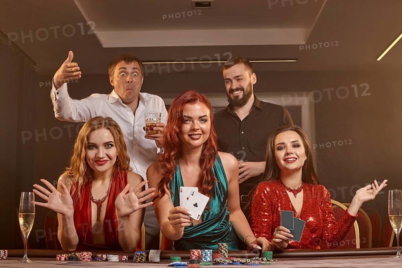 Happy friends are playing poker at casino. They are celebrating their win, smiling and posing at the table against a dark smoke background. Cards, chips, money, alcohol, fortune, gambling, entertainment concept