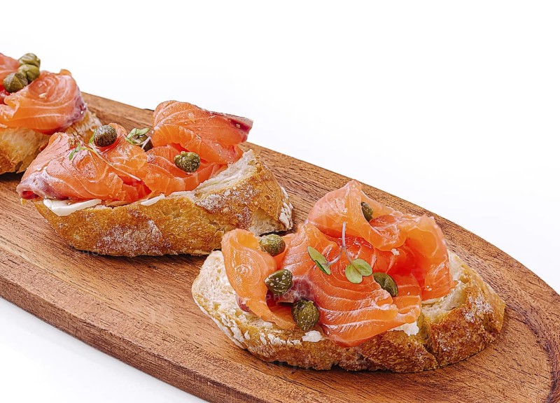 Food, Delicious canapés topped with smoked salmon and capers rest on a wooden platter. The vibrant colors of the salmon contrast nicely with the rustic bread, creating an appetizing sight
