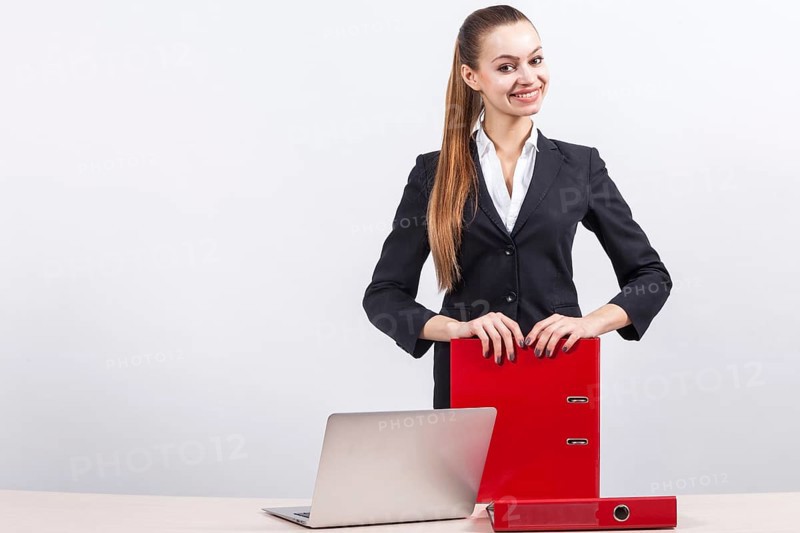 Attractive young caucasian businesswoman with ponytail in black suit stand near the office desk with laptop and hold red paper folders in hands, relies on folders Isolated on grey