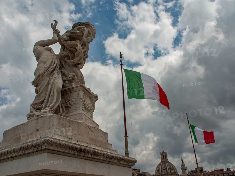 A statue and an Italian flag stand against a dramatic backdrop of clouds with ancient architecture in the background, rome, italy