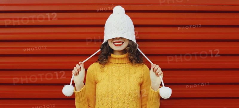 Winter christmas portrait of happy cheerful young woman smiling having fun in yellow sweater, white hat standing on red background