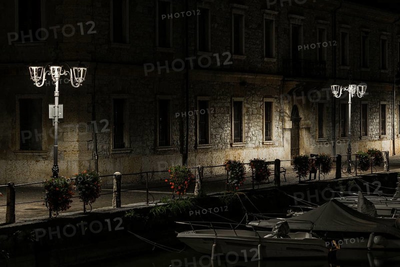 A canal view at night with moored boats, a stone building, and lampposts creating tranquil shadows, night photos taken at long exposure time taken in Peschiera del Garda, Italy Verona