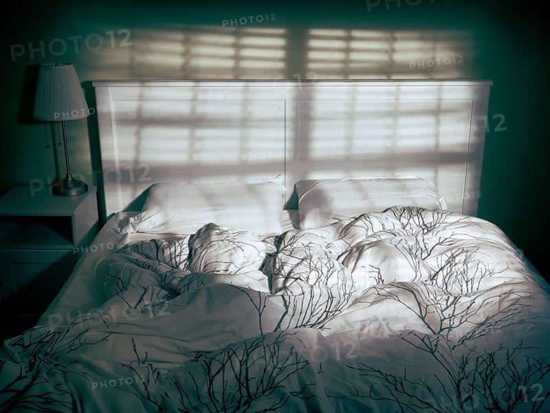 Unmade empty bed in dramatic dim light coming from a window through blinds