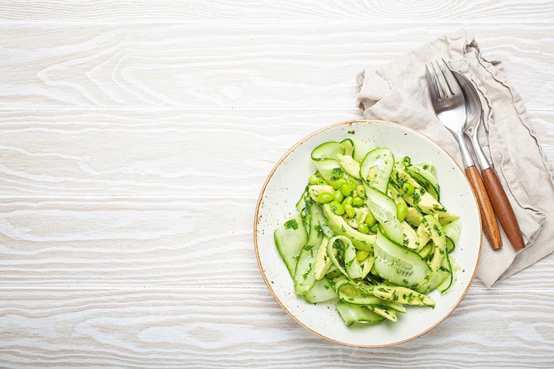 Healthy vegan green avocado salad bowl with sliced cucumbers, edamame beans, olive oil and herbs on ceramic plate top view on white wooden rustic table background. Space for text, food photography, food photography
