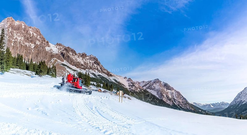 A sunny day of winter on the snowy pastures and the rocky peaks of the Austrian Alps mountains, with the roads being cleared by a snow groomer vehicle