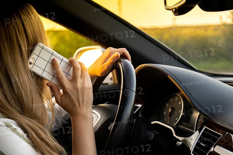 Young woman using mobile phone while driving car on highway road during sunset. Womandriver has accident calling with smartphone for help. Business woman busy driving concept