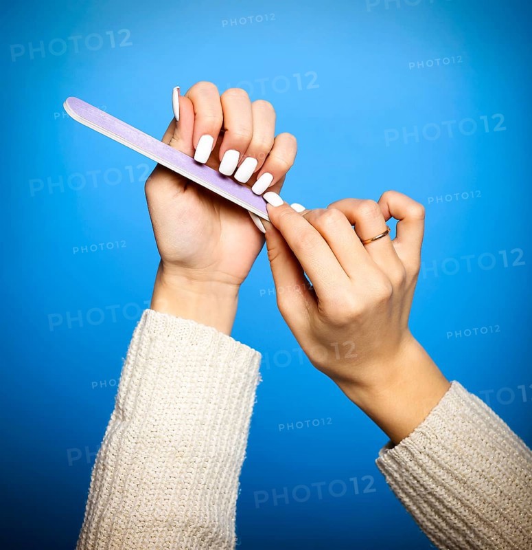 Faceless female in sweater filing long white nails doing manicure on blue background