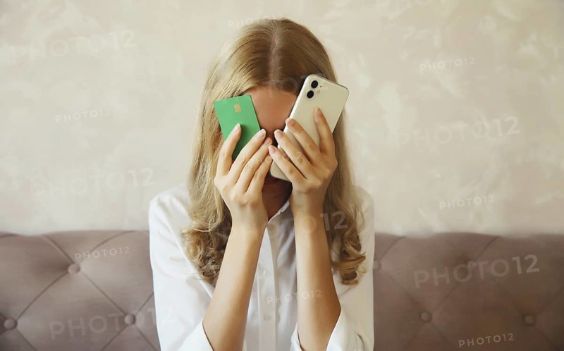 Desperate upset young woman holding mobile phone and bank card touching her head and covering face, internet fraud, problems with online payments or blocked credit card, sitting on the couch at home