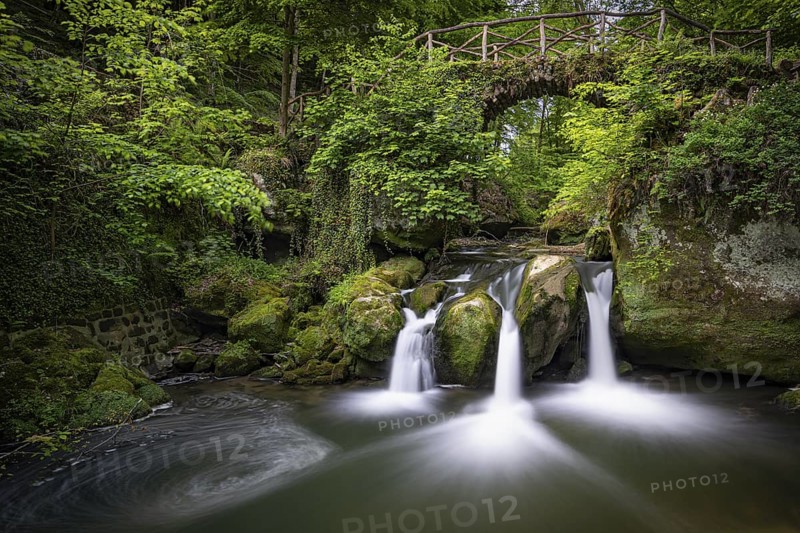 Three small waterfalls under a wooden bridge, surrounded by green vegetation, Mullerthal Region, Luxembourg Switzerland, Grand Duchy of Luxembourg